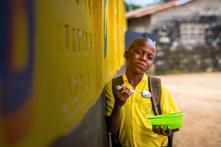 A boy leans against a yellow wall as he eats a bowl of food