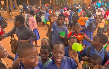 Children smiling and waving while holding mugs of food in a school playground.