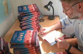 magnus macfarlane-barrow sitting at his desk signing a copy of his book, GIVE.