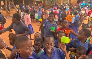 Children smiling and waving while holding mugs of food in a school playground.