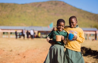 Two friends smiling together as they hold their mugs of food in school
