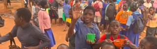 Group of children in a playground carrying mugs of food with one waving at the camera.