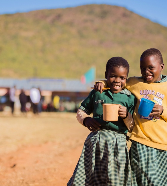 Two friends smiling together as they hold their mugs of food in school