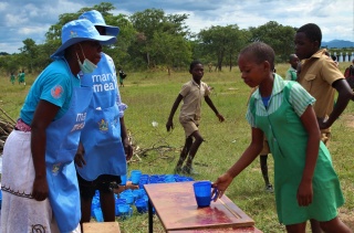 young girl approaching two Mary's Meals staff members at a table outdoors