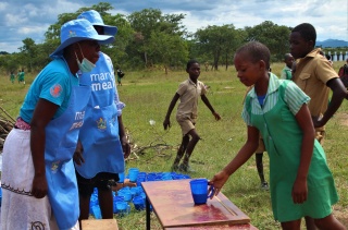 young girl approaching two Mary's Meals staff members at a table outdoors