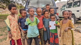 A group of children standing in front of some vehicles looking into the camera.