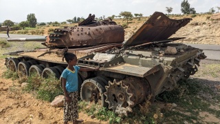 A child stands next to an abandoned tank next to a road.